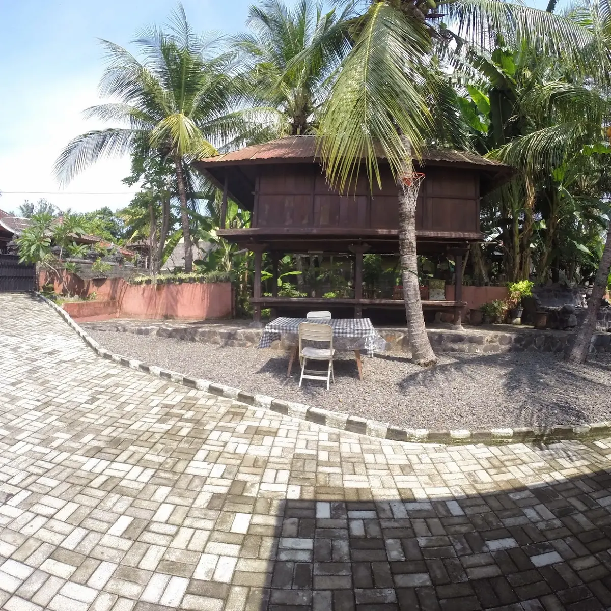 Paved courtyard with a lumbung pavilion and tall coconut palms at Homestay Dewata